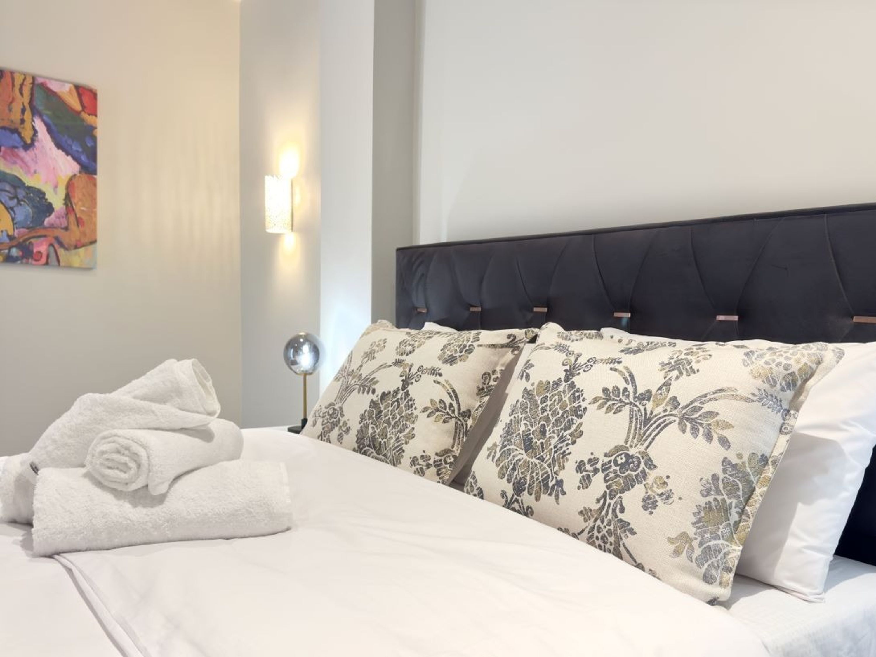 Una cama bien hecha con almohadillas estampadas, una toalla blanca enrollada y un cuadro en la pared.