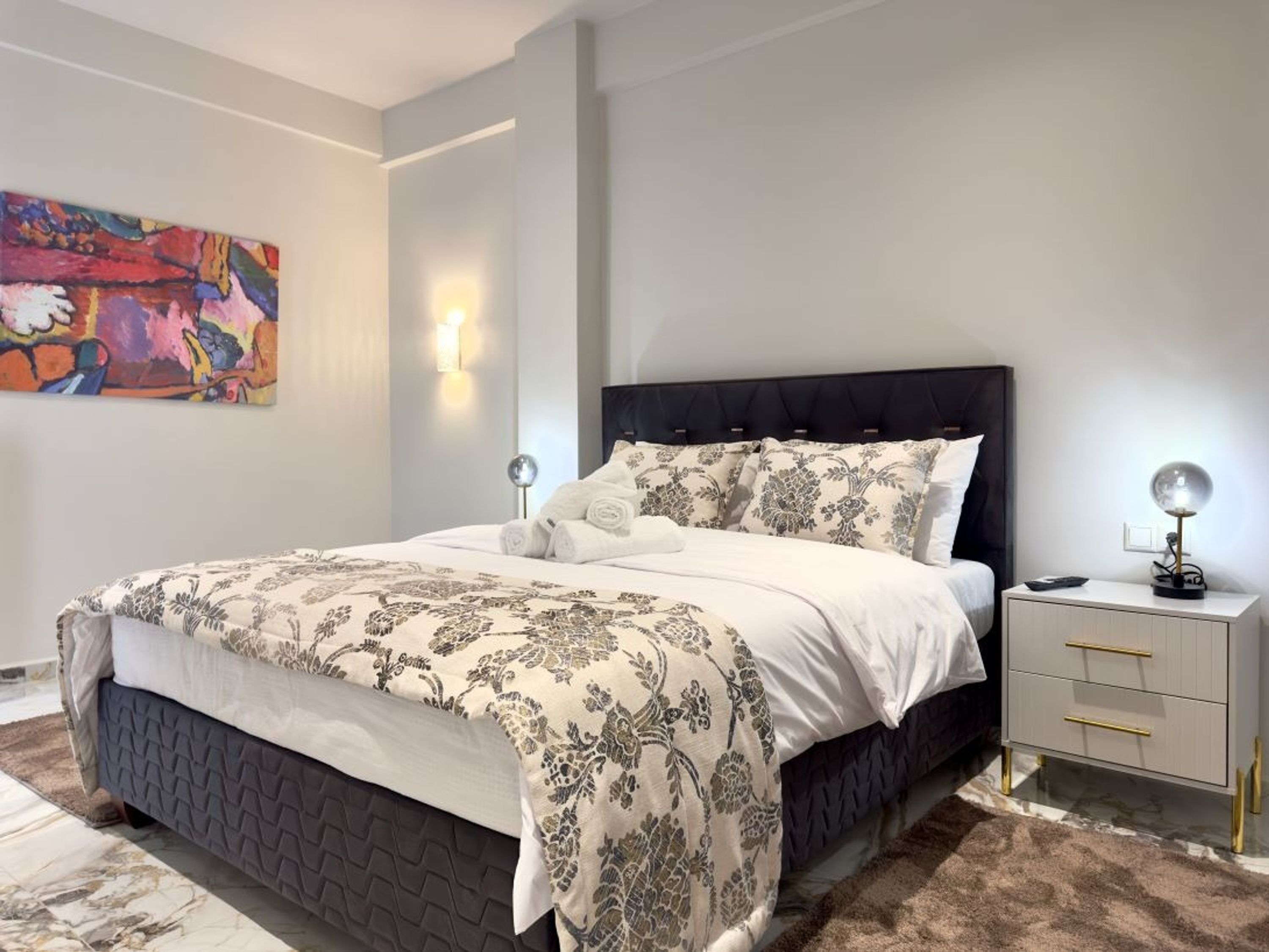 A modern bedroom with a large bed, a nightstand, a lamp, and a colorful abstract painting on the wall.