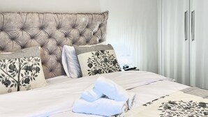 Cama bem arrumada com cabeceira estofada, almofadas estampadas com padrão floral e toalha dobrada sobre a cama.