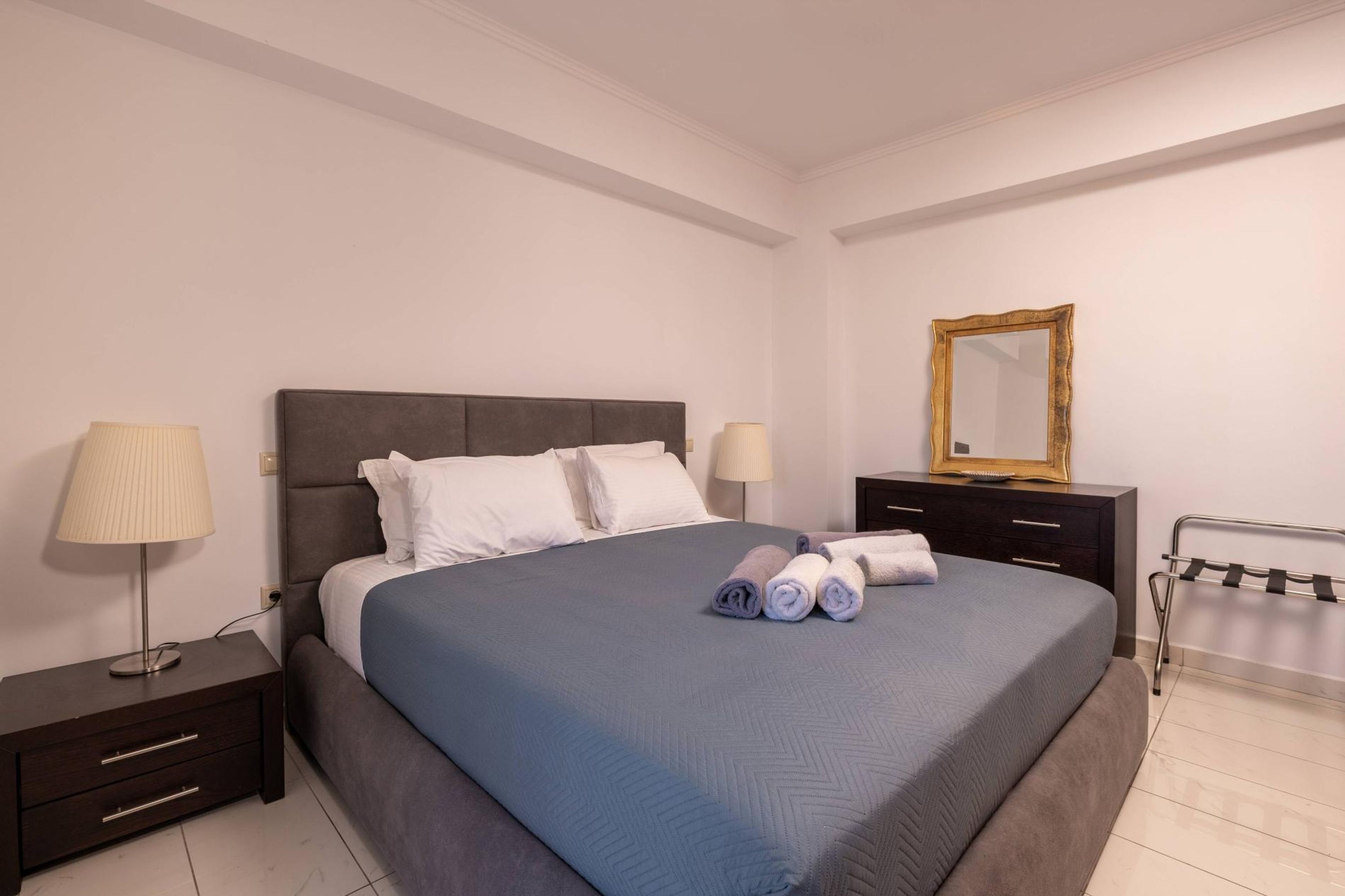 A neatly made bed with a blue bedspread, two bedside lamps, a dark wooden nightstand with drawers, a black metal rack, and a gold-framed mirror on the wall.