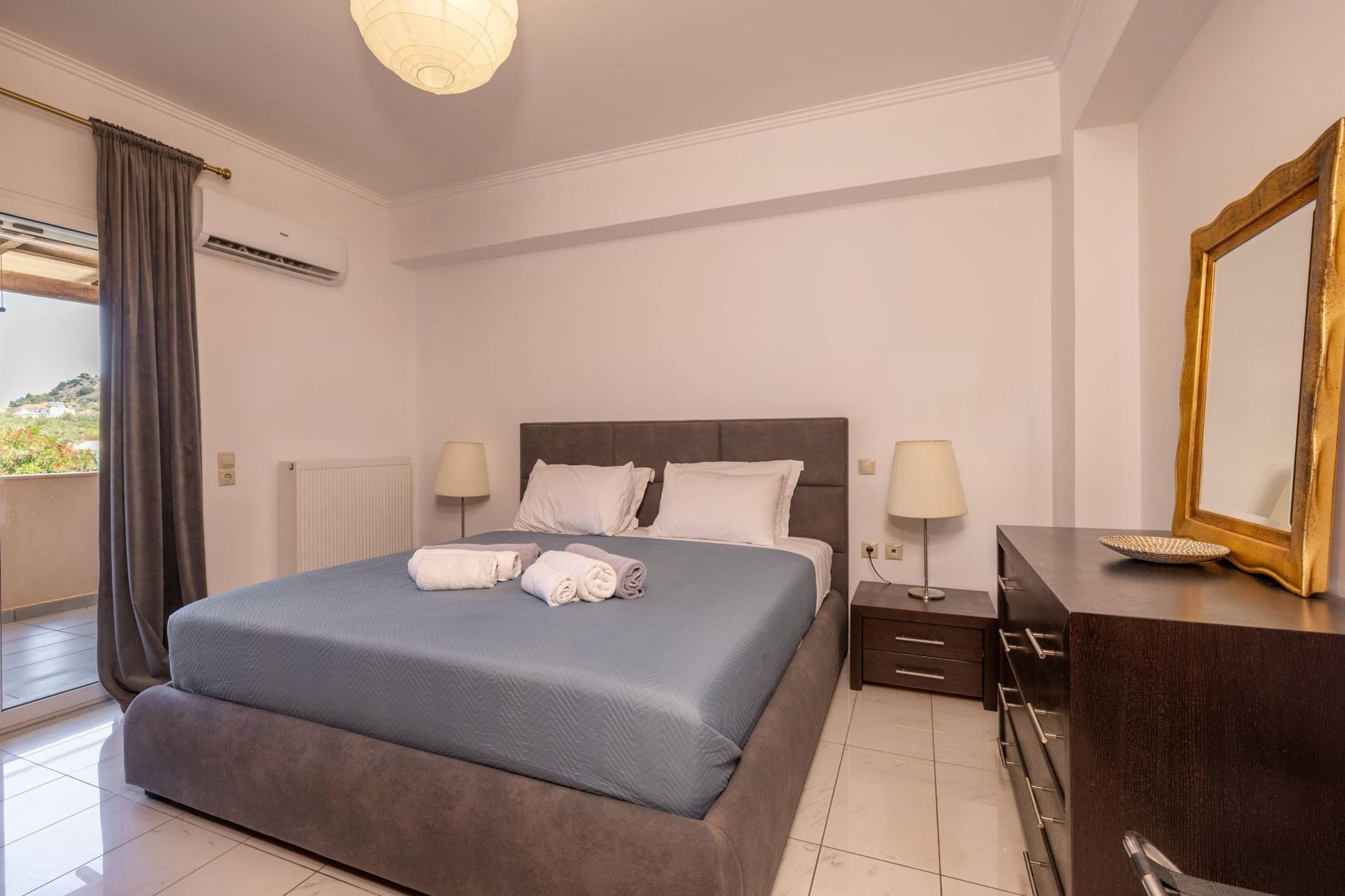 Een net opgemaakt bed met kussens en opgevouwen handdoeken, een nachtkastje met een lamp, een dressoir met een spiegel en een raam met gordijnen.