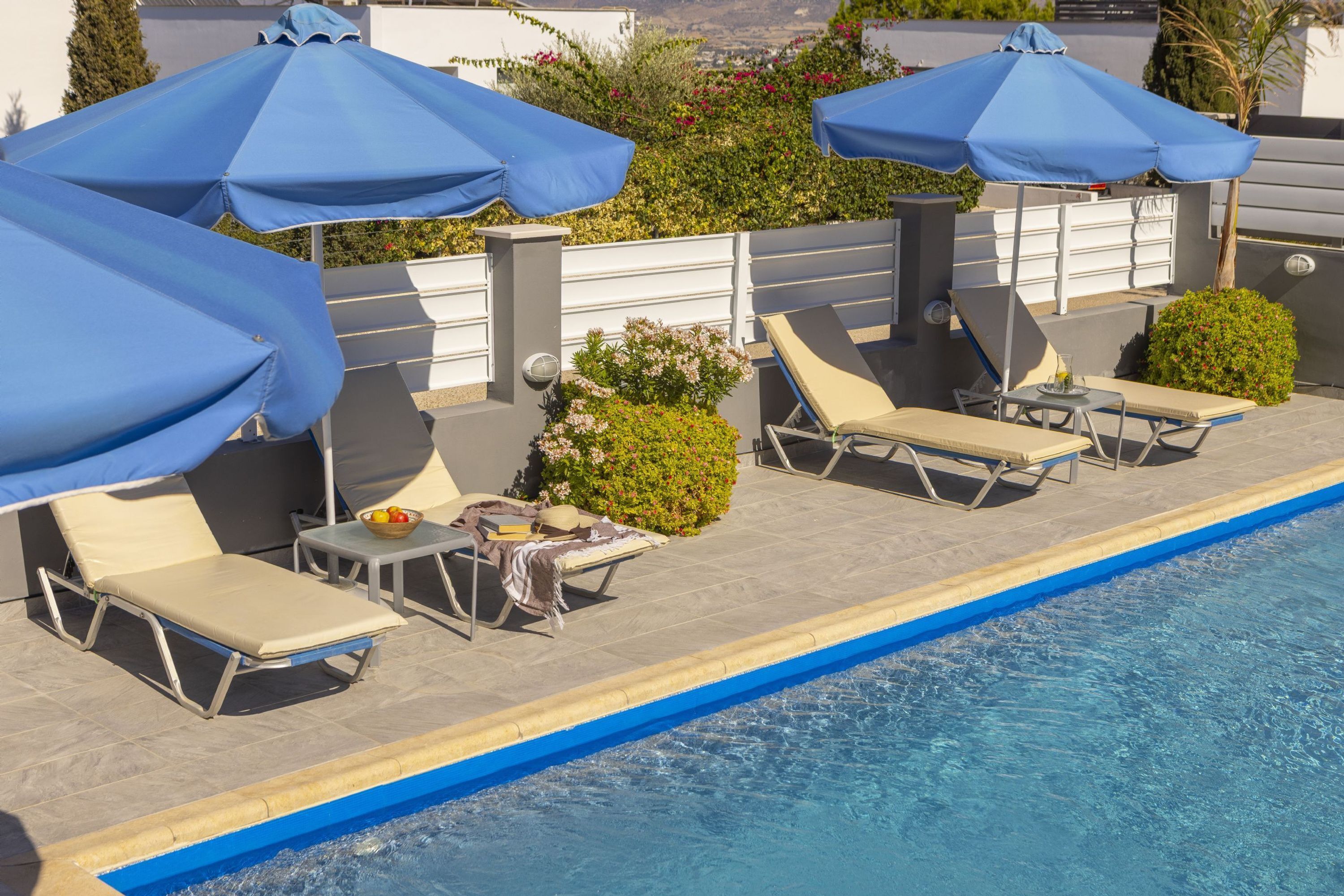 A poolside area with lounge chairs and umbrellas, a small table with a bowl of fruit, and a view of greenery and buildings in the background.