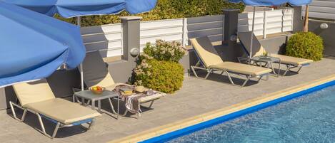 A poolside area with lounge chairs and umbrellas, a small table with a bowl of fruit, and a view of greenery and buildings in the background.