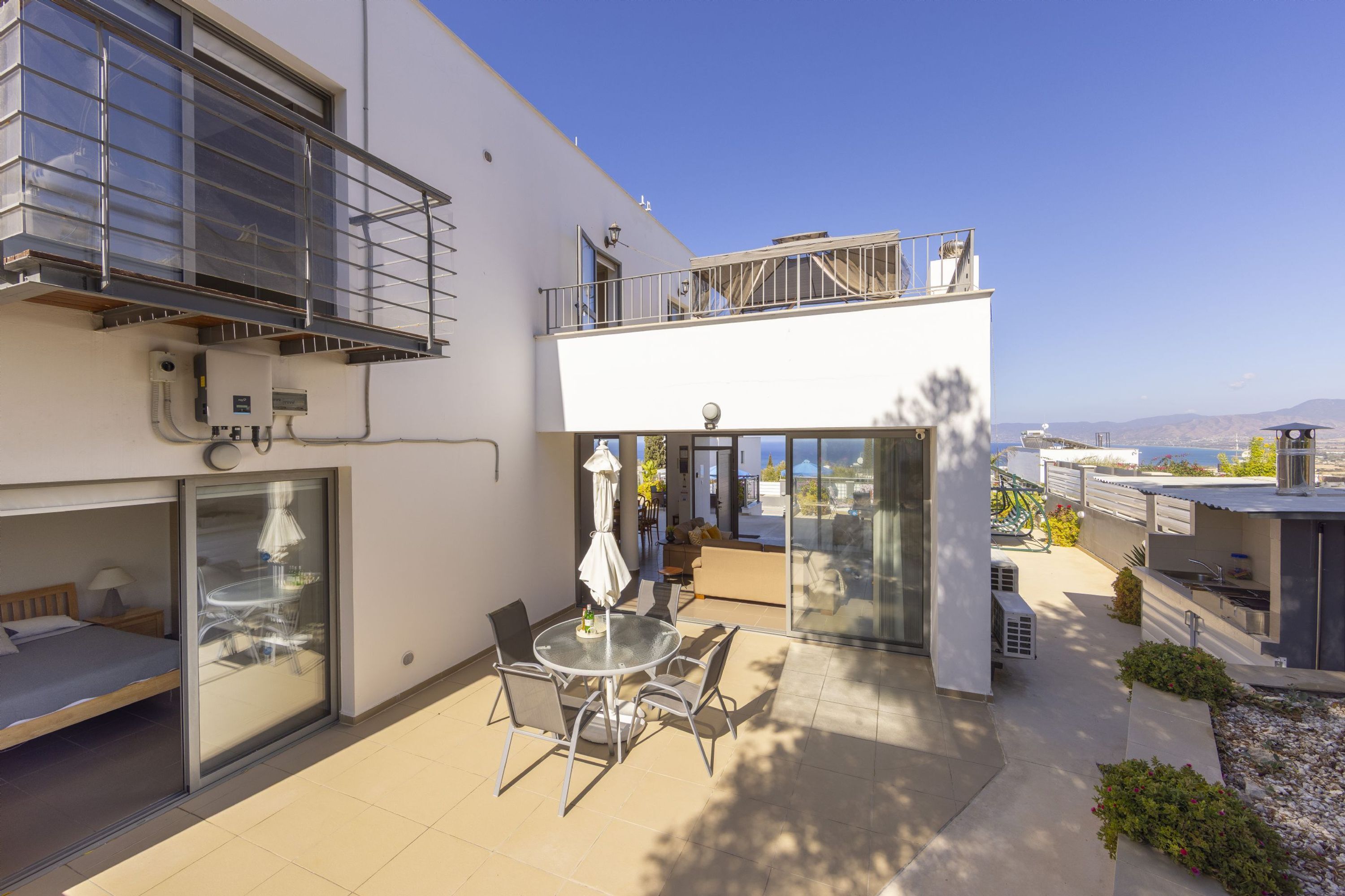 A modern two-story house with a balcony, a glass door leading to a patio with a table and chairs, and a view of the landscape.