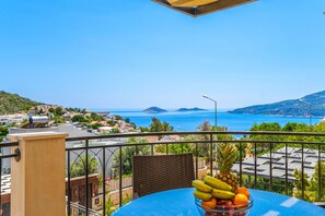 A balcony with a table and a bowl of fruit, overlooking a coastal town with buildings and a clear blue sky. - Villa Vanilla in Kalkan (Kalkan)