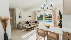 A modern living room with a sofa, coffee table, and dining table. There is a large window with a view of a swimming pool. - Aiolia Villa in Rhodes (Rhodes)