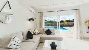 A modern living room with a beige sectional sofa, a glass coffee table, and a view of a swimming pool through large glass doors. - Aiolia Villa in Rhodes (Rhodes)