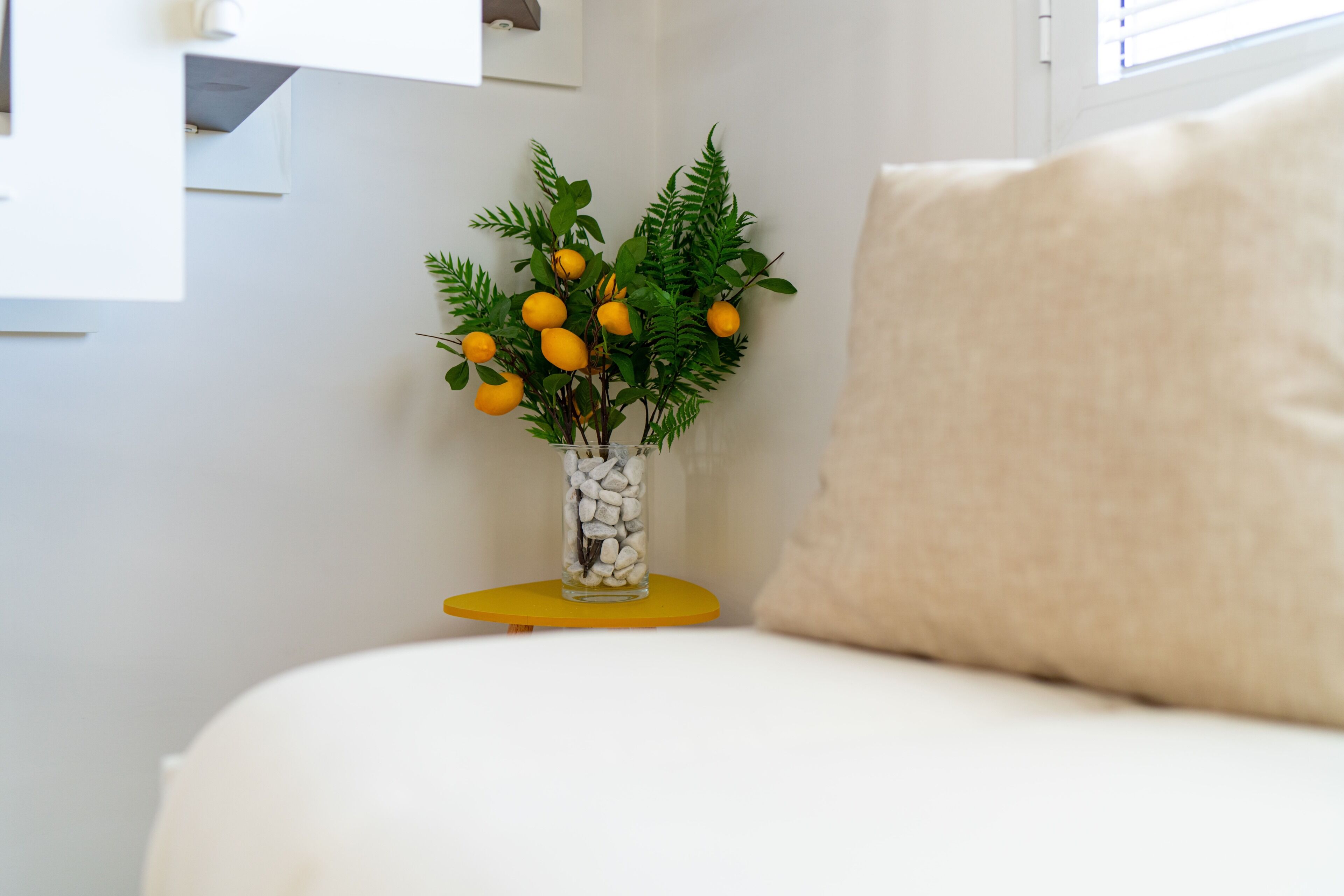 Un sofá blanco con un cojín beige, una mesita auxiliar amarilla de tamaño pequeño con un jarrón de vidrio que contiene una planta con hojas verdes y frutos amarillos, y una pared blanca con una ventana.