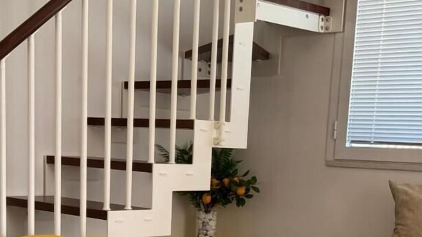 A room with a yellow chair, a bed with a beige cover, a small table with a vase, and a staircase with white railings.