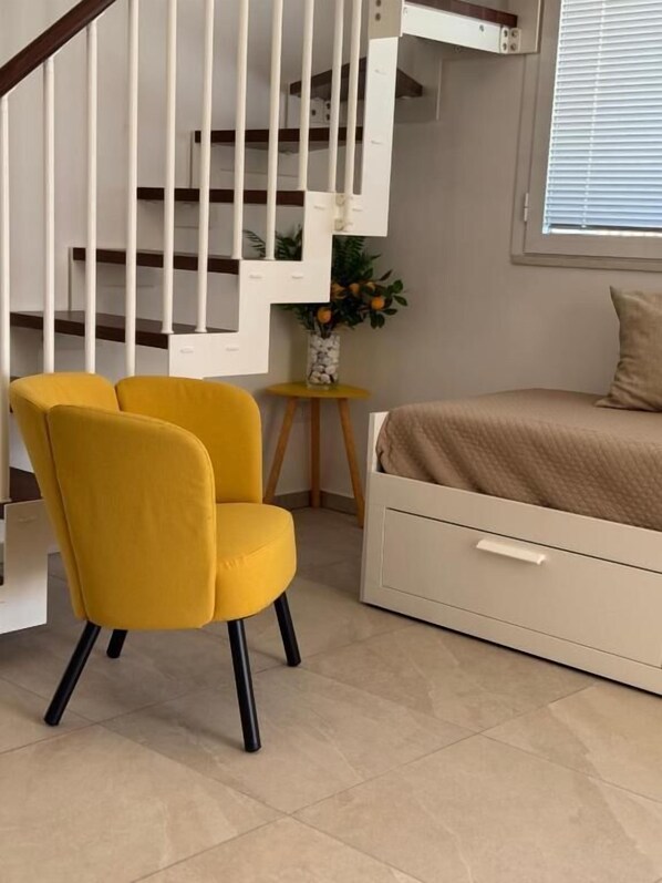 A room with a yellow chair, a bed with a beige cover, a small table with a vase, and a staircase with white railings.