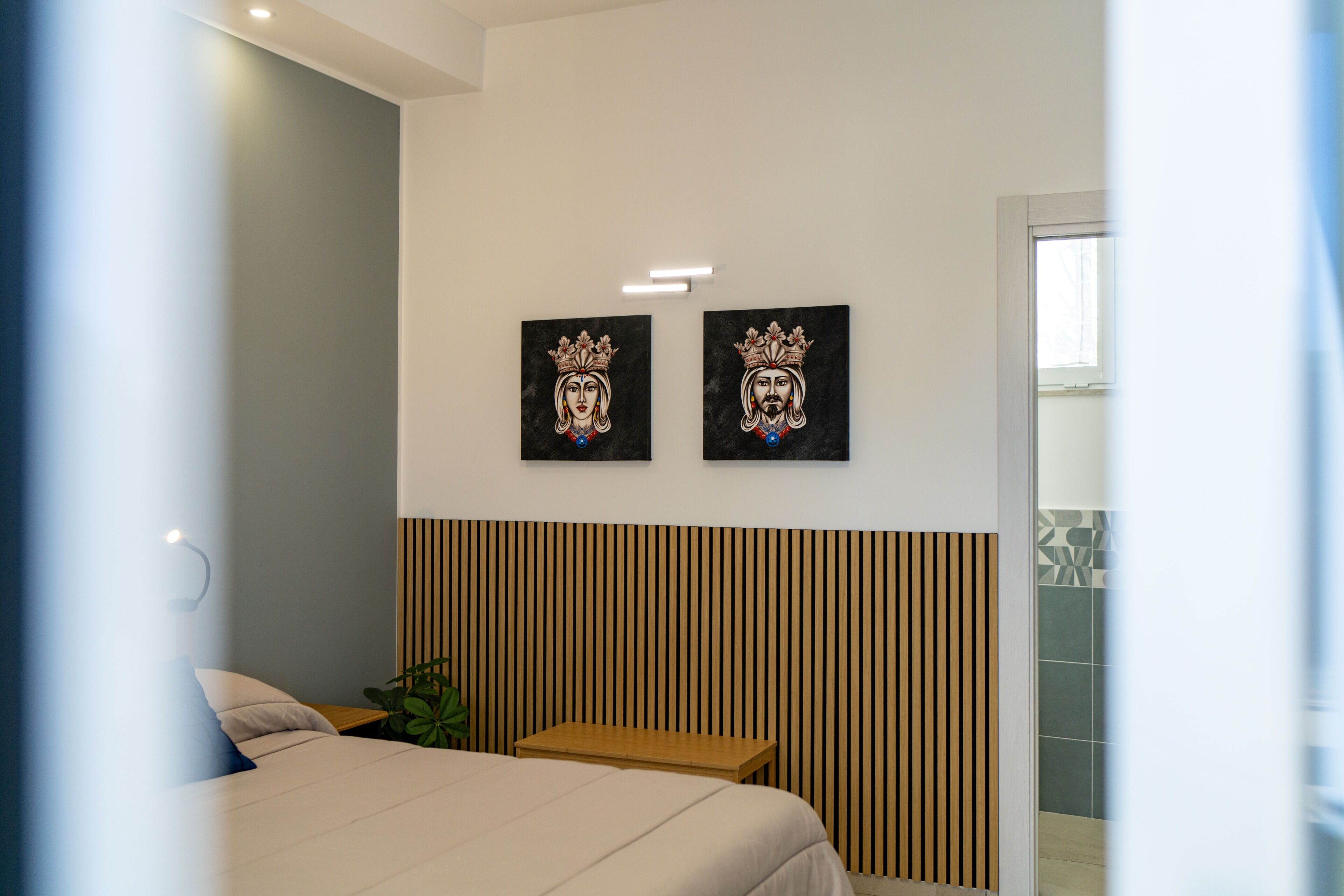A hotel room with a bed, a small table, two wall-mounted artworks, and a wooden slat headboard.