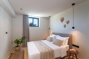 A modern bedroom with a large bed, a potted plant, a small table, and a window with curtains. - Massari Hills- Villa Finis Terrae 2 (Rhodes)