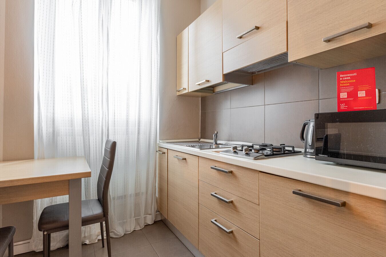 Une cuisine compacte équipée d’un four à micro-ondes, d’un grille-pain et d’une bouilloire. On y trouve une table à manger avec une chaise, ainsi qu’un panneau d’accueil rouge accroché au mur.