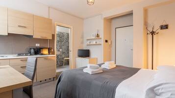 A compact hotel room with a kitchenette, a bed with white linens, a desk with a chair, and a television.