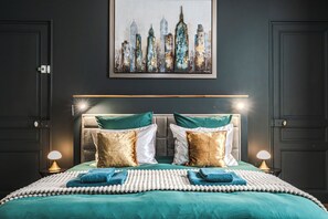 A bedroom with a cityscape painting, a bed with teal bedding, two bedside lamps, and a headboard with a light fixture.