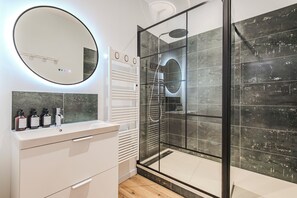 Ein modernes Badezimmer mit einer Duschkabine aus Glas, einem runden Spiegel und einem weißen Waschtisch mit Pflegeprodukten.