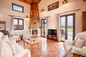 A spacious living room with a stone fireplace, comfortable seating, a flat-screen TV, and large windows offering a view of the outdoors. - Villa Levante in Kefallonia (Kefallonia)