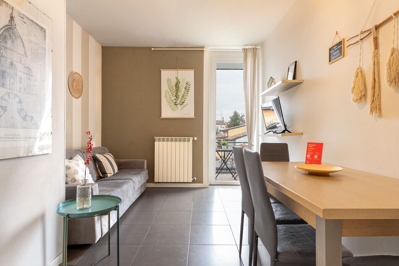 A modern living room with a sofa, a dining table, and chairs. There is a radiator, a framed artwork on the wall, and a view of the outside through an open door.
