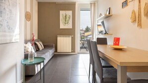 Ein modernes Wohnzimmer mit einer Couch, einem Esstisch und Stühlen. Ein Heizkörper, ein gerahmtes Kunstwerk an der Wand und ein Blick ins Freie durch eine geöffnete Tür sind ebenfalls zu sehen.