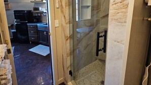 A modern bathroom with a glass shower enclosure, marble walls, and a visible kitchen area in the background. - The Silo On 16 (Graham)