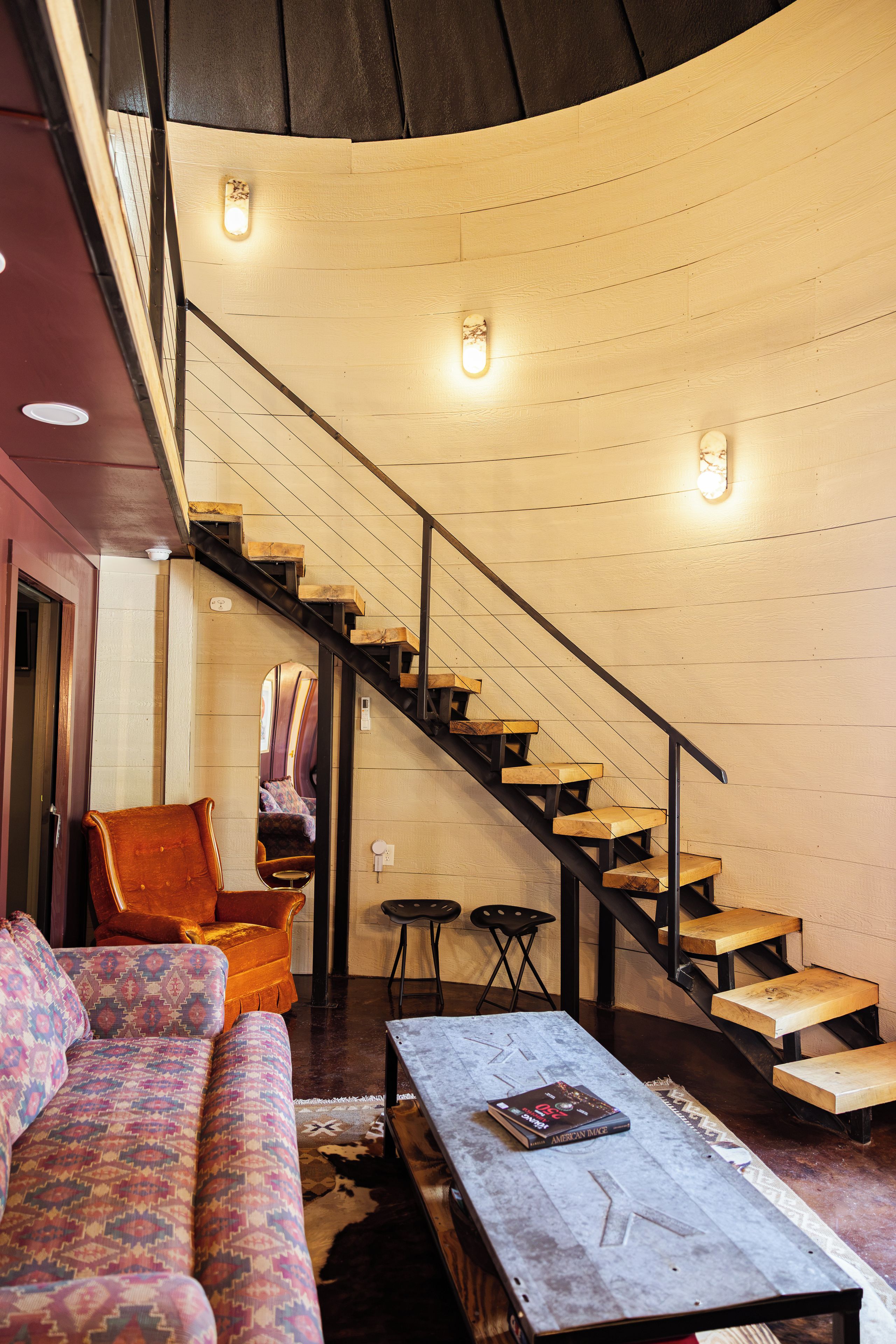 A cozy interior with a curved staircase, a sofa, a coffee table, and a mirror reflecting a chair and a bed.