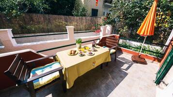 Eine Terrasse mit einem für zwei Personen gedeckten Tisch, umgeben von Holzstühlen, einer gelben Tischdecke und einem orangefarbenen Sonnenschirm.