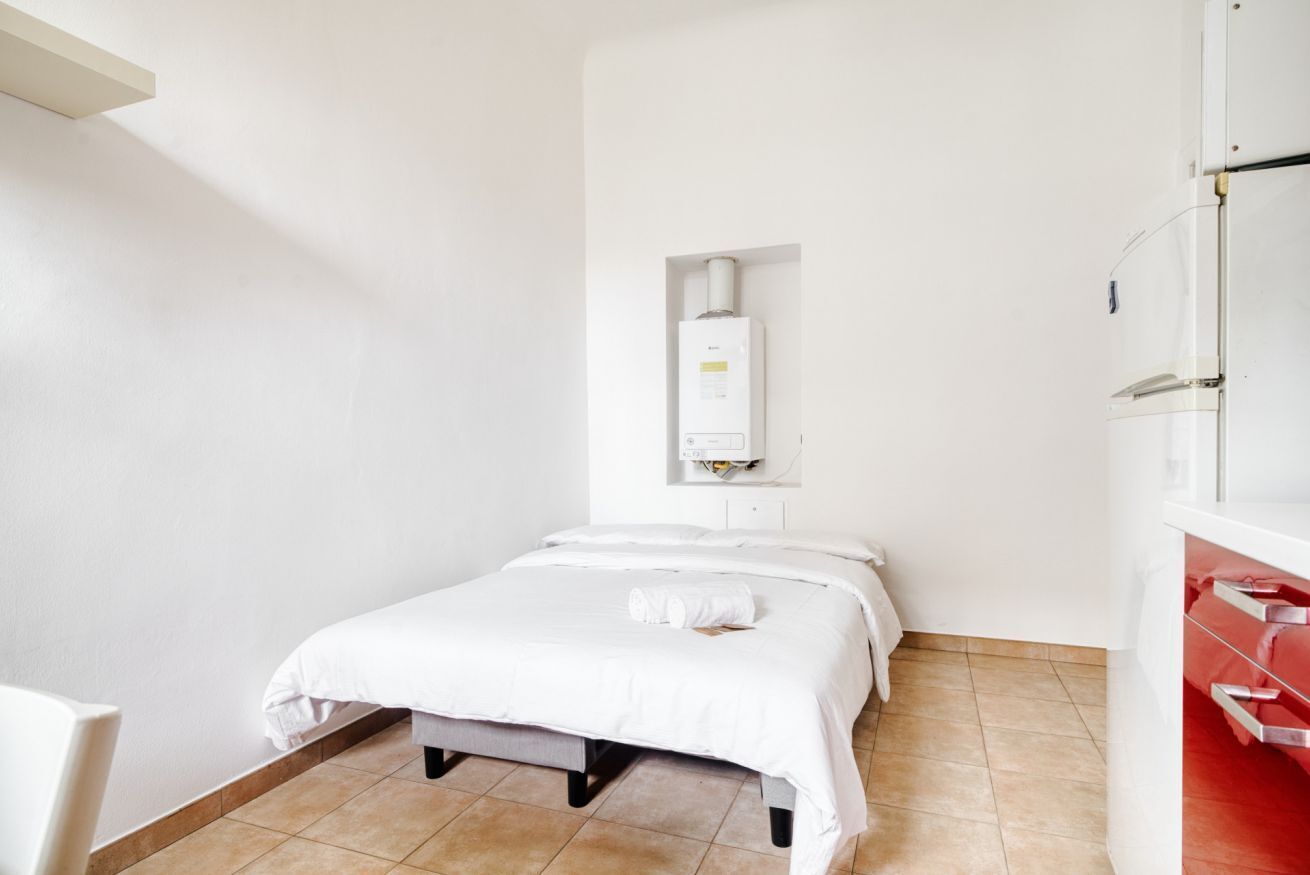 A single-bed room with a white bed, a small bedside table, a wall-mounted heater, and a red cabinet.