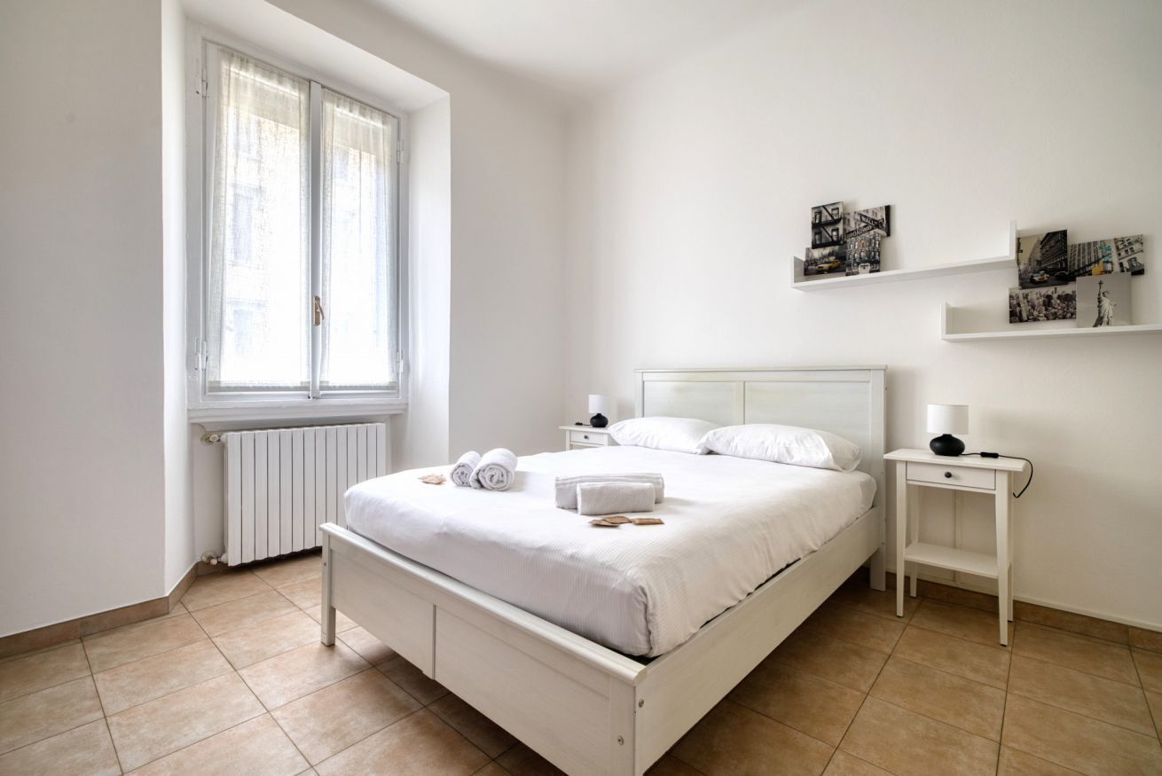Une chambre à coucher comprenant un lit, deux tables de chevet, une fenêtre avec des rideaux et des étagères murales décorées de tableaux.
