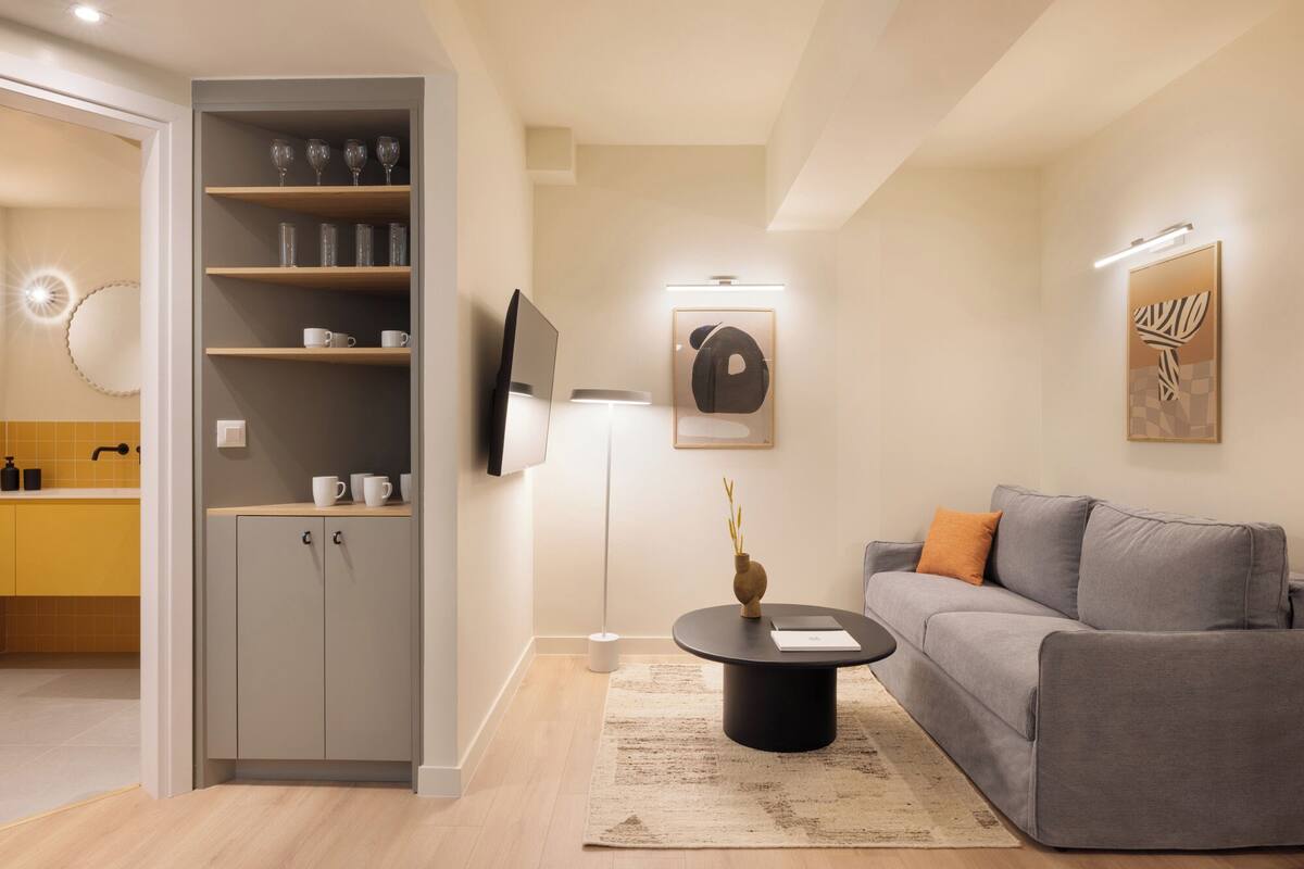 A modern living room with a grey sofa, a coffee table, a bookshelf with glasses and cups, a flat-screen TV, and a framed artwork on the wall.
