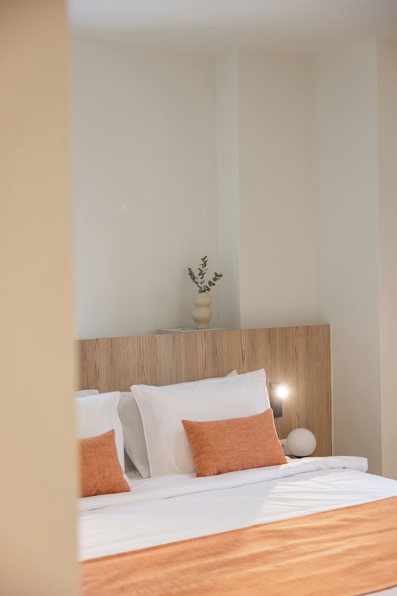 Ein Hotelzimmer mit einem hölzernen Kopfteil, einem Bett mit weißer Bettwäsche und orangefarbenen Kissen, einer Vase mit Grünpflanzen und einer runden Wandleuchte.