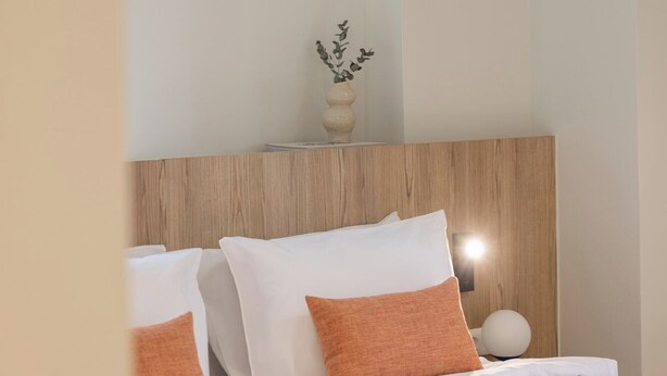 Une chambre d’hôtel avec une tête de lit en bois, un lit avec une literie blanche et des oreillers orange, un vase contenant des feuilles vertes et un luminaire rond fixé au mur.