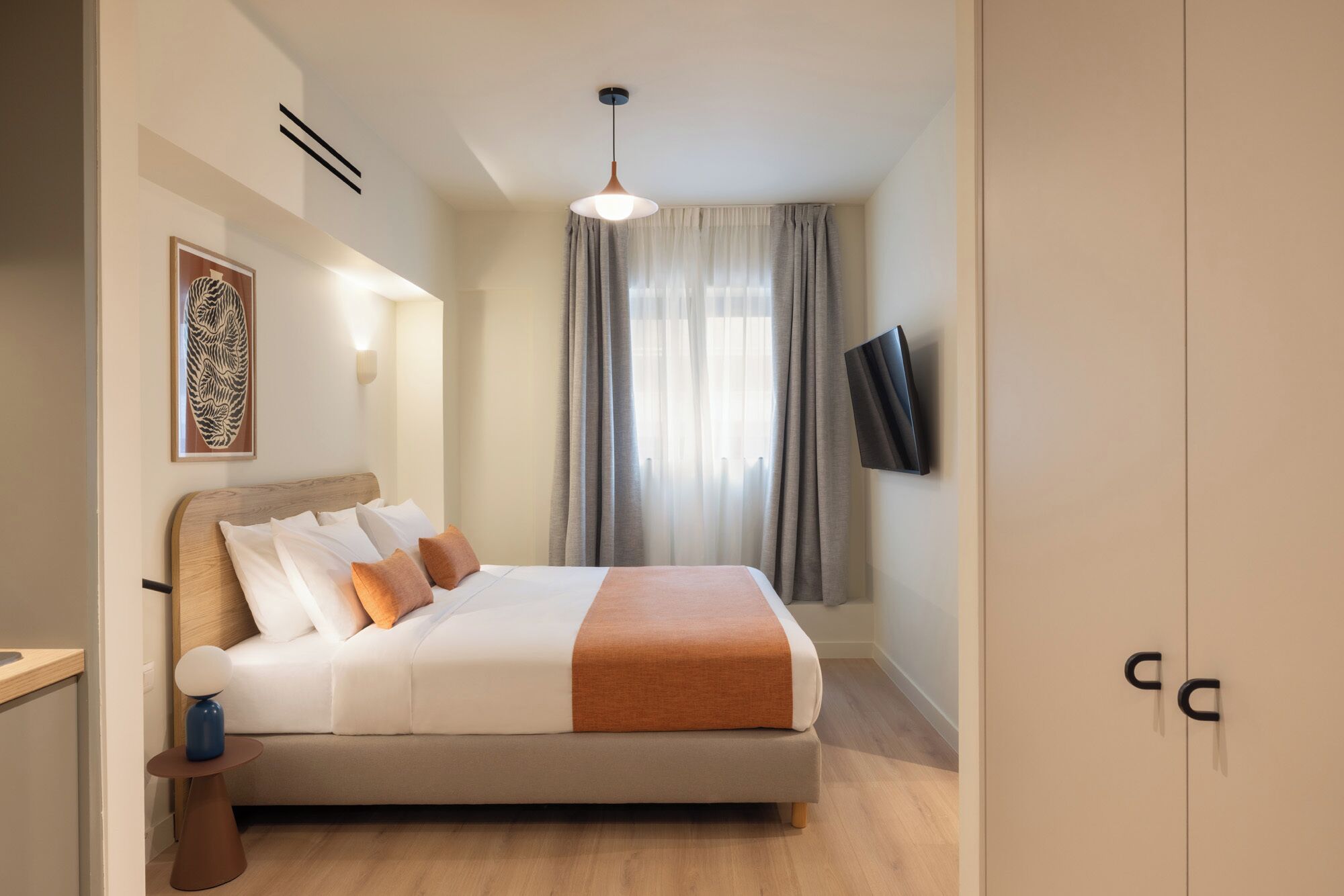 A hotel room with a bed, a bedside table with a lamp, a wall-mounted TV, and a large window with curtains.