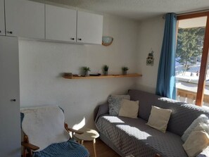 Ein Zimmer mit einer Couch, einem Sessel, einem kleinen Tisch, einem Regal mit Pflanzen und einem Fenster mit Blick.