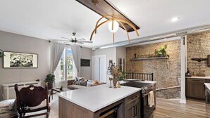 A modern kitchen with a central island, a dining area with a high ceiling fan, and a living room with a sofa and a fireplace.