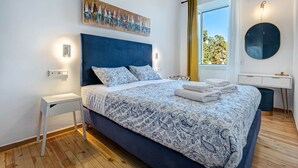 Een slaapkamer met een groot bed, een nachtkastje, een wandlamp, een schilderij, een raam met gordijnen en een ronde spiegel.