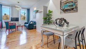 Een moderne woonkamer met een glazen eettafel, stoelen met een blauwgroen en zwart schaakpatroon, een blauwe bank en een groot raam met gordijnen.
