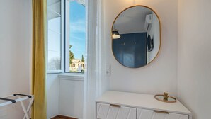 Een kamer met een witte kaptafel, een ronde spiegel, een raam met gele gordijnen en uitzicht op gebouwen.