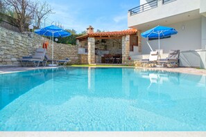 Ein Schwimmbecken mit Liegestühlen und Sonnenschirmen, ein Grillplatz aus Stein und eine Steinsäule.