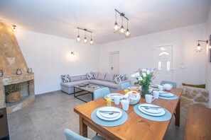 Ein Essbereich mit Holztisch, gedeckt für das Frühstück, einem Kamin aus Stein und einem modernen Wohnbereich mit Sofa und Leuchten.