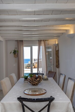 A dining area with a table set for six, a hanging plant, and a view of the sea through a glass door. - Οrmos seaside villa A with private pool (Tinos)