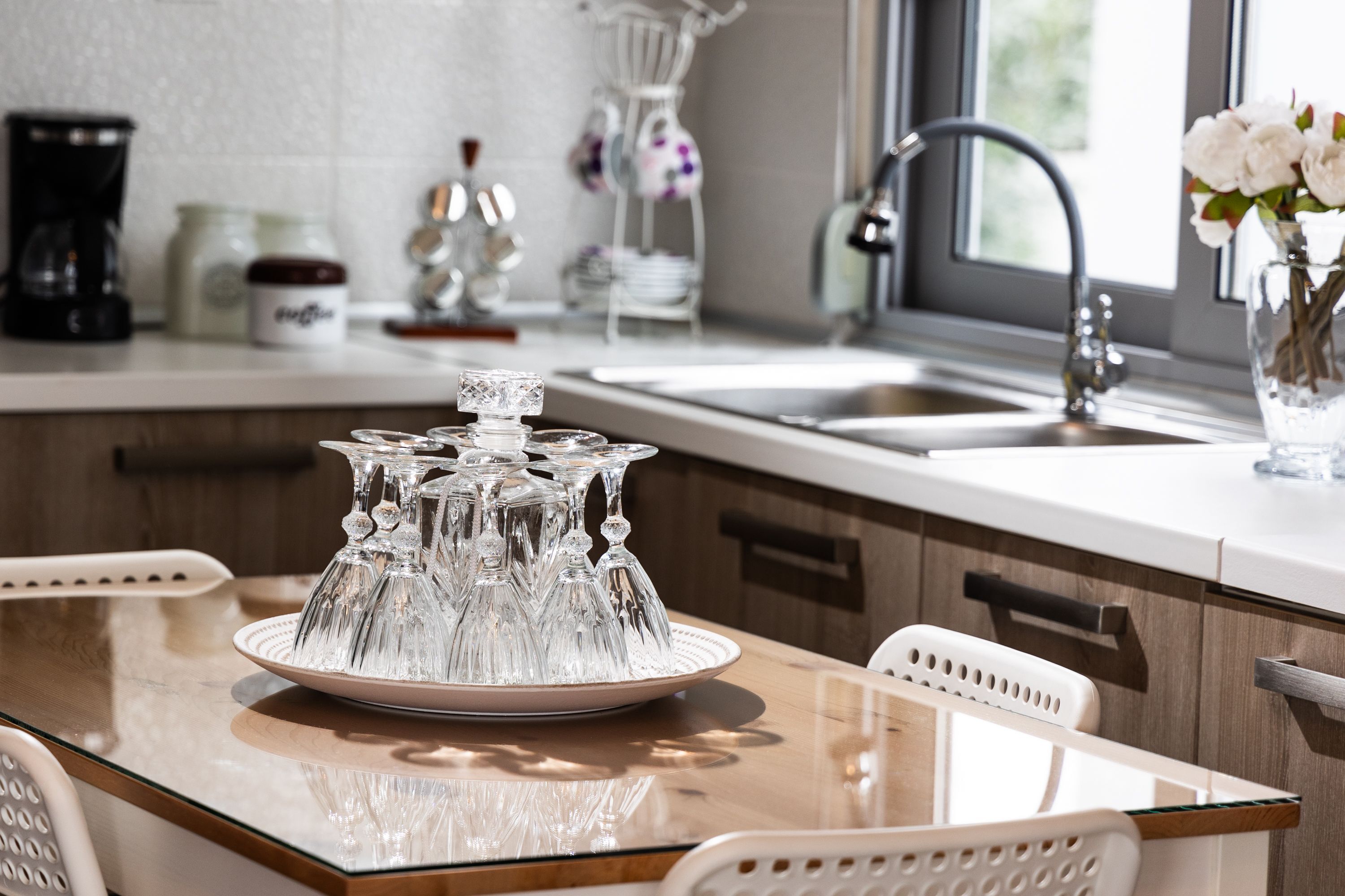 Een keuken met een glazen tafel, witte stoelen, een spoelbak en een vaas met bloemen.
