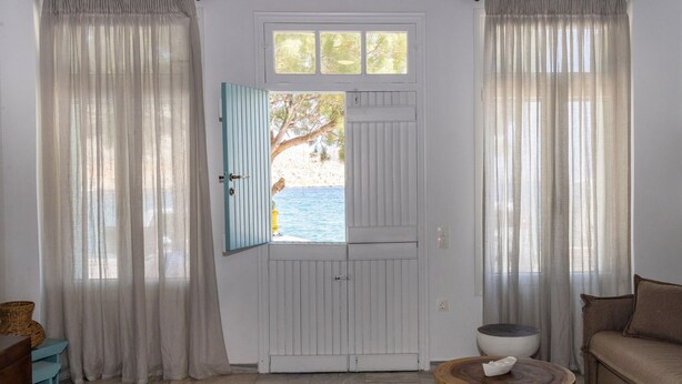 A room with a view of the sea through an open door, a wooden table with a bowl, a sofa, and a small round table. - Aelios Cycladic house (Ormos Panormou)