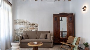 A cozy living room with a stone wall, a wooden cabinet, a sofa, a rattan chair, and a round wooden table. - Aelios Cycladic house (Ormos Panormou)