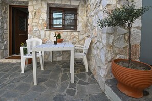 Un bâtiment en pierre avec une porte en bois, une fenêtre avec des barreaux en métal, une table blanche avec des chaises et une plante en pot.