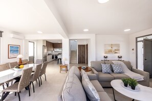 A modern living room with a sofa, dining table, and chairs. There is a kitchen in the background with a refrigerator and a sink. - Melissoula Villa in Neo Chorio (Neo Chorio)