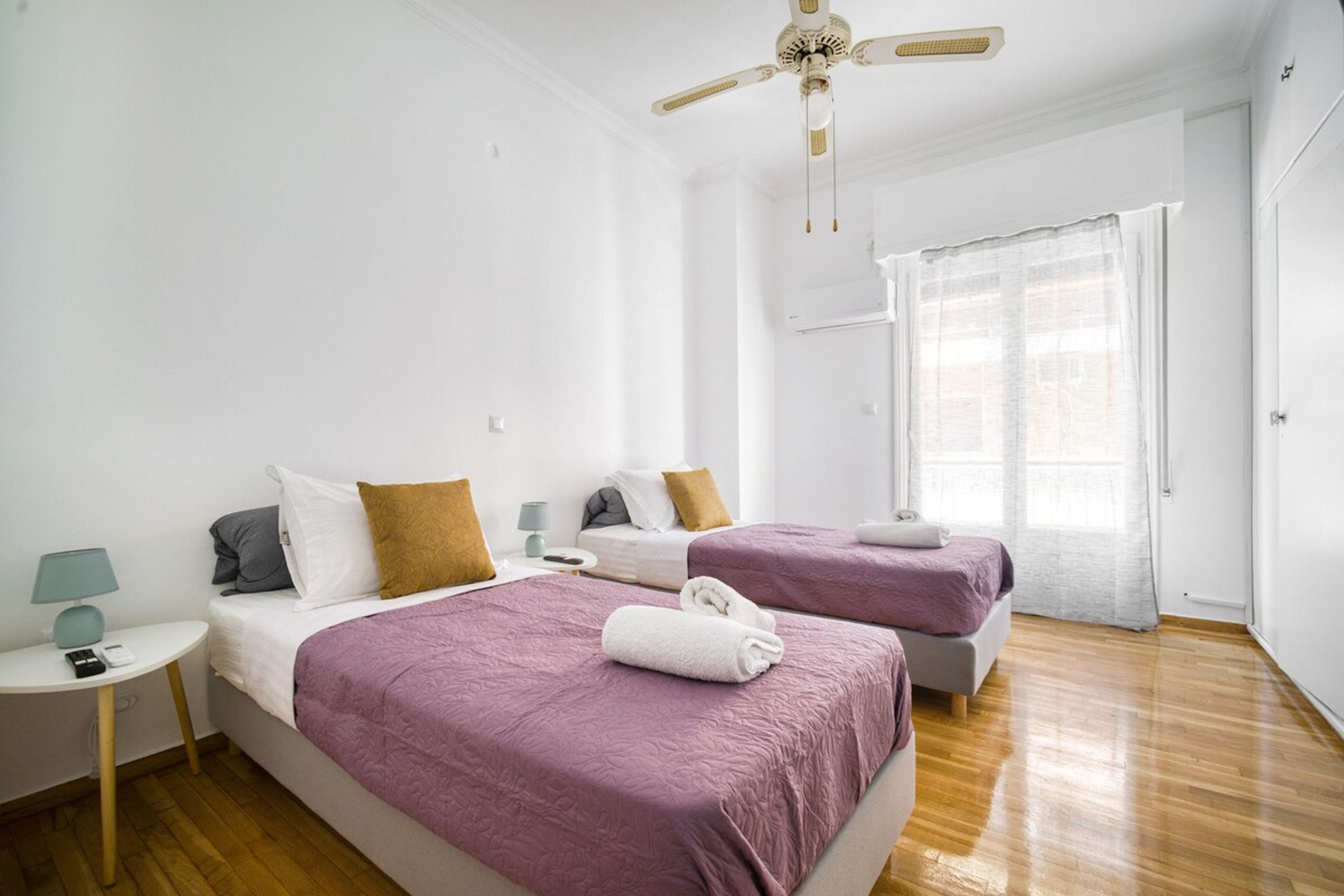 A hotel room with two beds, a ceiling fan, a window with sheer curtains, a bedside table with a lamp, and a wooden floor.