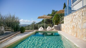 En swimmingpool med klart vand, omgivet af stenvægge og gelænder i glas. To beige parasoller og en trappe op til et højere niveau.