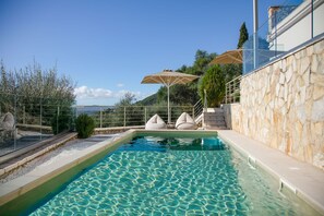 Ein Pool mit klarem Wasser, umgeben von Steinmauern und einer Glasreling. Zwei beigefarbene Sonnenschirme und eine Treppe, die zu einer erhöhten Ebene führen.