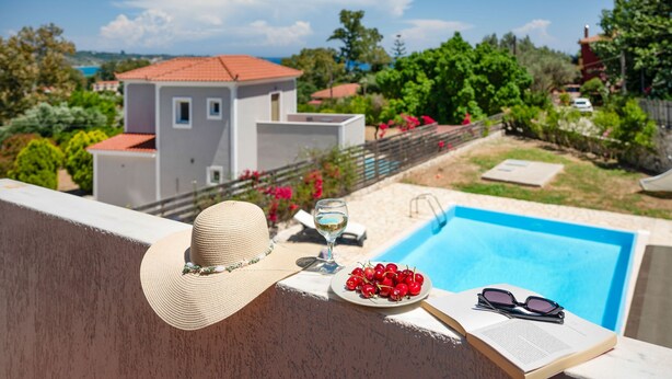 Blick auf einen Pool mit einem weißen Gebäude, einem Hut, einer Schale mit Kirschen, einem Weinglas und einer Sonnenbrille auf einem Tisch.