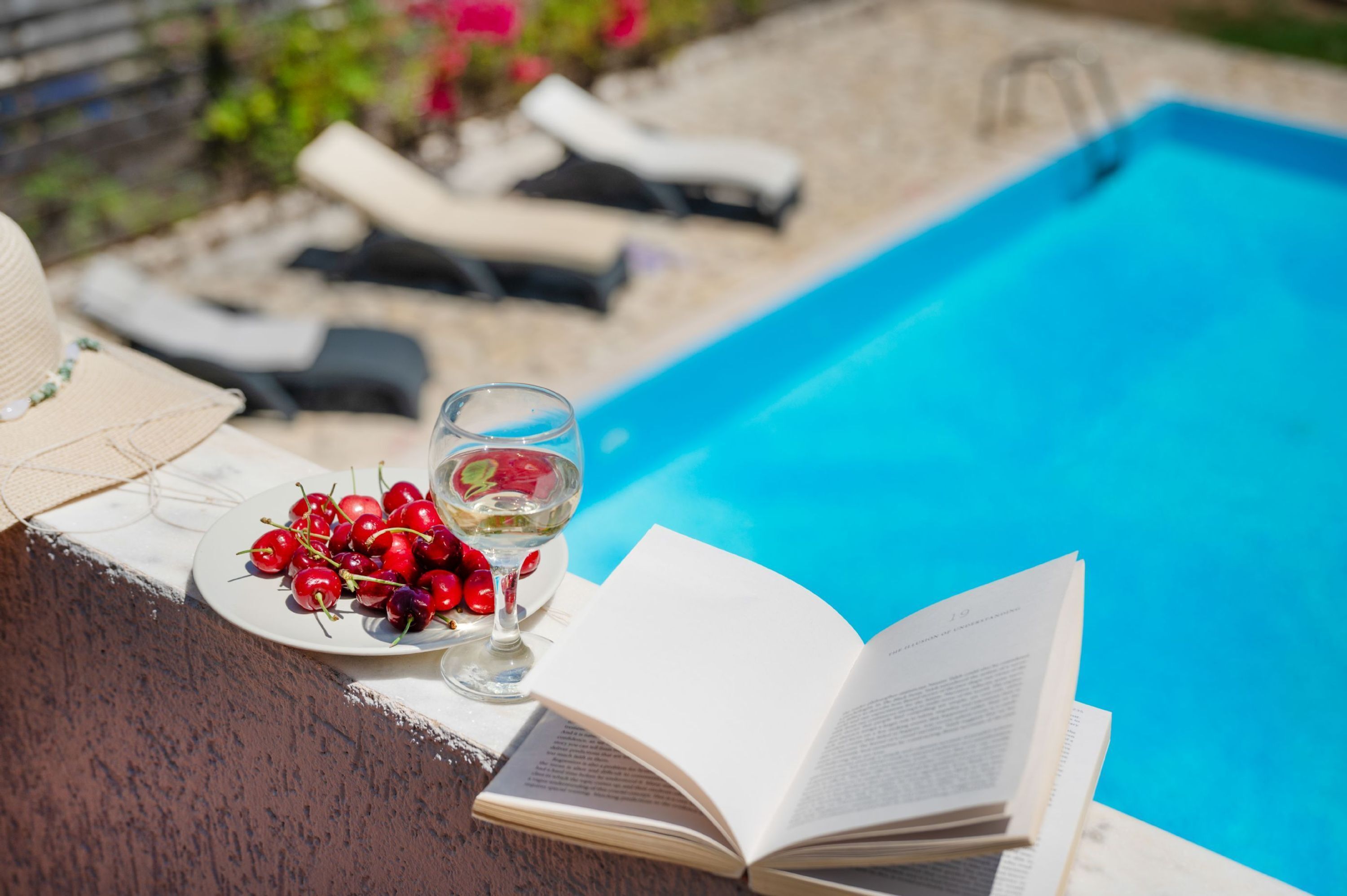 Ein Poolbereich mit Buch, Glas Weißwein und einer Schale Kirschen.
